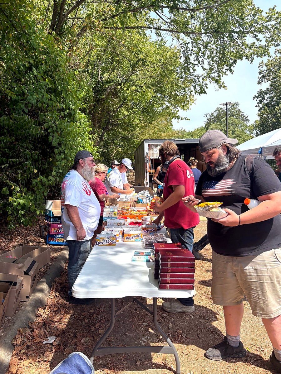 Neighbors filling plates along a table of food and supplies under the trees